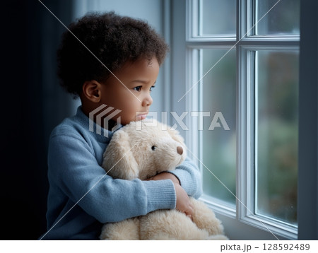 African young child with teddy bear looking out window in thoughtful pose. 128592489