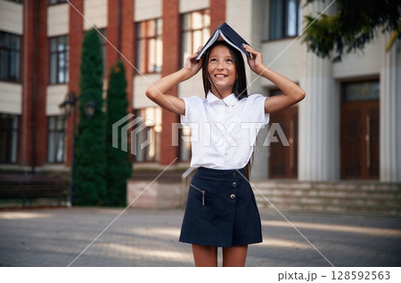 Playing with book that is over the head. School girl in uniform is outdoors near the building 128592563