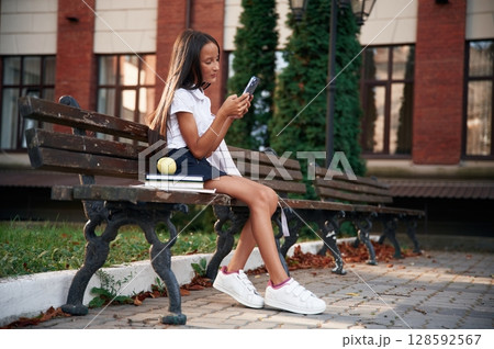 Sitting on the bench with smartphone. School girl in uniform is outdoors near the building Sitting on the bench with smartphone. School girl in uniform is outdoors near the building 128592567
