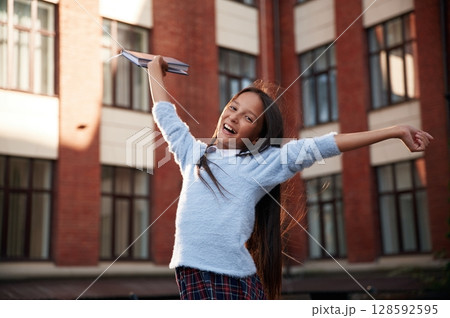 Cheerful, celebrating success. School girl in uniform is outdoors near the building Cheerful, celebrating success. School girl in uniform is outdoors near the building 128592595