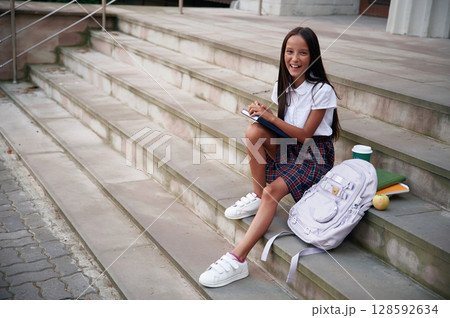 Having free time, sitting on the stairs. School girl in uniform is outdoors near the building 128592634