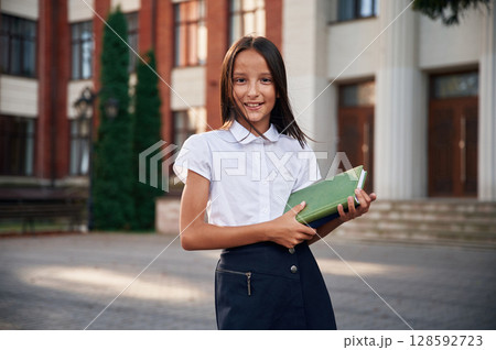 Standing, front view, holding books. School girl in uniform is outdoors near the building 128592723