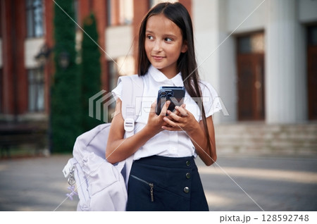 Holding smartphone. School girl in uniform is outdoors near the building 128592748