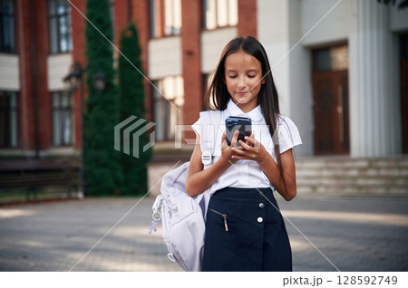 Phone in hands. School girl in uniform is outdoors near the building Phone in hands. School girl in uniform is outdoors near the building 128592749