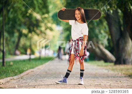 Holding the skateboard in the hand, on the shoulder. Happy little girl is outdoors Holding the skateboard in the hand, on the shoulder. Happy little girl is outdoors 128593165