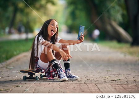 Showing tongue, making selfie. Happy little girl with skateboard outdoors 128593176