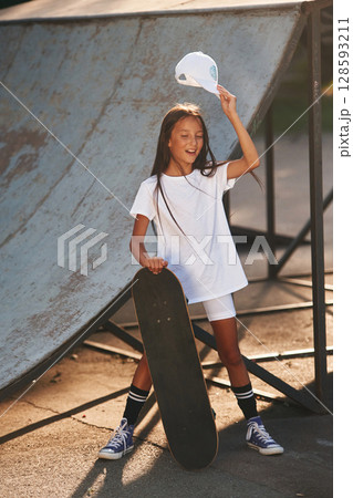Standing with skateboard. Happy little girl is outdoors Standing with skateboard. Happy little girl is outdoors 128593211