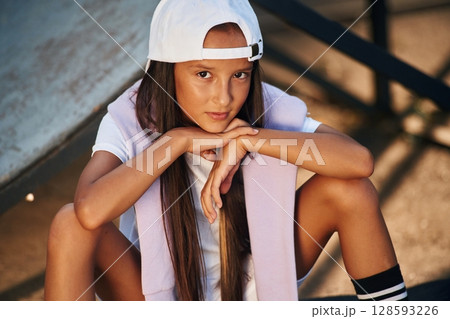 Sitting and looking at the camera. Young girl in casual clothes is in the public park outdoors Sitting and looking at the camera. Young girl in casual clothes is in the public park outdoors 128593226