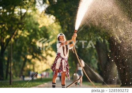 Plays with water as it pours from a hose. Happy little girl with skateboard outdoors Plays with water as it pours from a hose. Happy little girl with skateboard outdoors 128593272