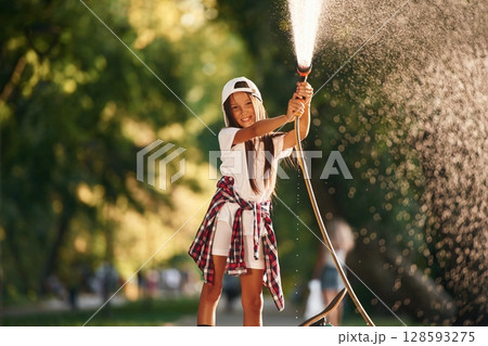Plays with water as it pours from a hose. Happy little girl with skateboard outdoors Plays with water as it pours from a hose. Happy little girl with skateboard outdoors 128593275