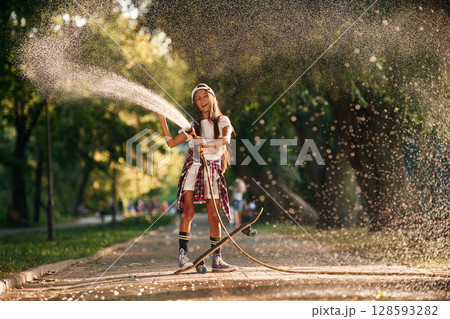 Pouring water from a hose. Happy little girl with skateboard outdoors Pouring water from a hose. Happy little girl with skateboard outdoors 128593282