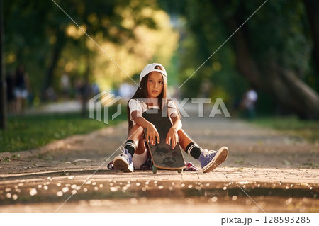 Green trees are in the background, on the path. Happy little girl with skateboard outdoors 128593285