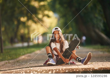 Green trees are in the background, on the path. Happy little girl with skateboard outdoors 128593286