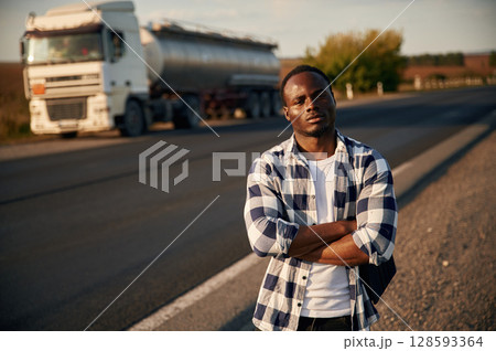 Serious look. With arms crossed. Black man is standing on the road with truck on it 128593364