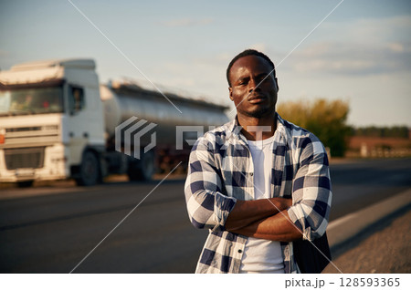 Serious look. With arms crossed. Black man is standing on the road with truck on it 128593365