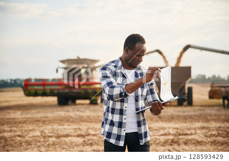 Opposite agricultural machinery. Beautiful African American man is in the agricultural field Opposite agricultural machinery. Beautiful African American man is in the agricultural field 128593429