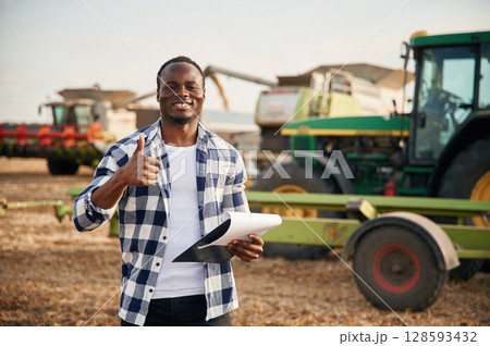 Opposite agricultural machinery. Beautiful African American man is in the agricultural field 128593432