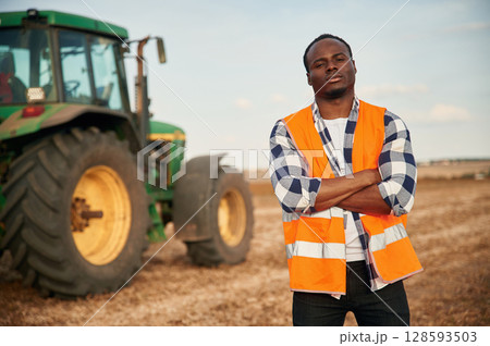 Near the tractor in orange uniform. Beautiful African American man is in the agricultural field 128593503