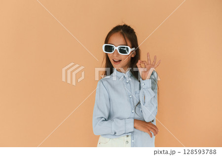 Okay hand gesture, in sunglasses. Cute young girl is in the studio against background Okay hand gesture, in sunglasses. Cute young girl is in the studio against background 128593788