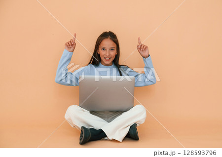 Pointing to the top. Sitting with laptop. Cute young girl is in the studio against background Pointing to the top. Sitting with laptop. Cute young girl is in the studio against background 128593796