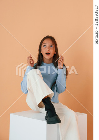 Sitting on cube with fingers crossed. Cute young girl is in the studio against background 128593817