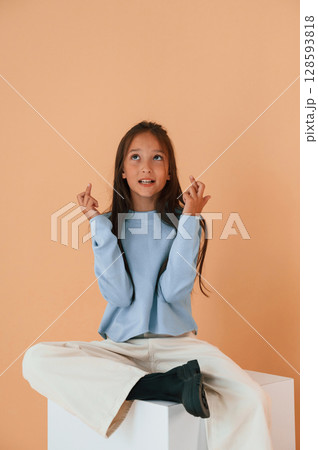 Sitting on cube with fingers crossed. Cute young girl is in the studio against background 128593818