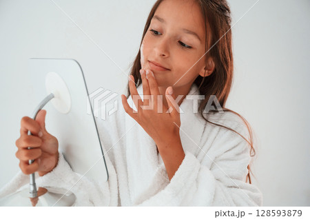 Using cream on face, holding mirror. Conception of beauty and self care. Young girl is in the studio 128593879