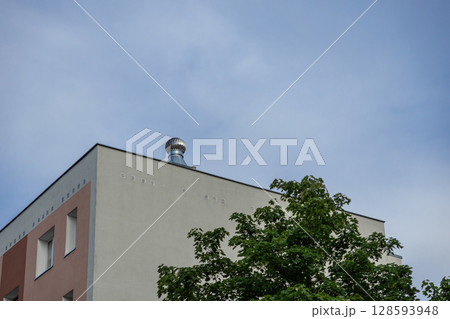 Modern building corner showcasing a functional rooftop ventilation system and lush green tree against a clear sky, highlighting sustainable urban infrastructure development Modern building corner showcasing a functional rooftop ventilation system and lush green tree against a clear sky, highlighting sustainable urban infrastructure development 128593948