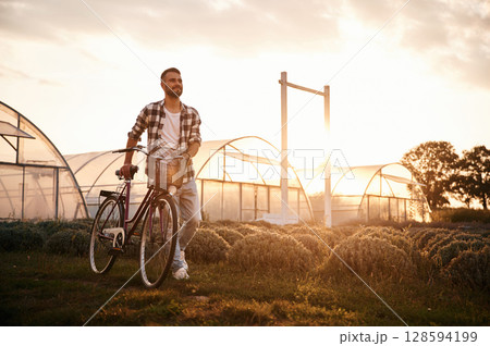 Against sunlight. Handsome man in casual clothes is with bicycle on the agricultural field near greenhouse 128594199