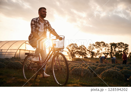 Riding bicycle. Handsome man in casual clothes is on the agricultural field near greenhouse Riding bicycle. Handsome man in casual clothes is on the agricultural field near greenhouse 128594202