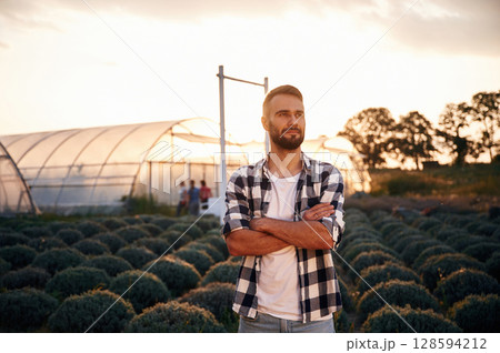 Serious young man on agricultural field is standing with arms crossed near the greenhouse that is behind Serious young man on agricultural field is standing with arms crossed near the greenhouse that is behind 128594212