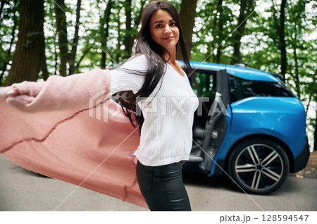 Enjoying nature and free time, active. Cheerful beautiful woman is near blue car outdoors Enjoying nature and free time, active. Cheerful beautiful woman is near blue car outdoors 128594547