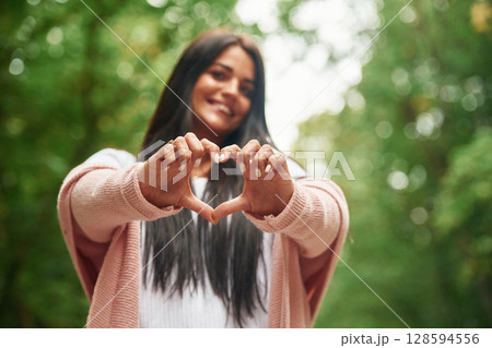 Showing heart gesture by hands. Cheerful beautiful woman is standing outdoors, green leaves at background Showing heart gesture by hands. Cheerful beautiful woman is standing outdoors, green leaves at background 128594556