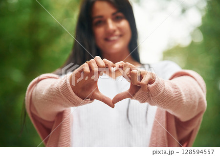 Showing heart gesture by hands. Cheerful beautiful woman is standing outdoors, green leaves at background Showing heart gesture by hands. Cheerful beautiful woman is standing outdoors, green leaves at background 128594557