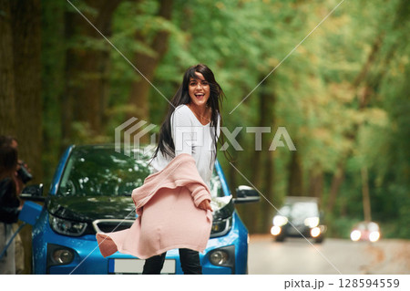 Black automobile behind. Cheerful beautiful woman is near blue car outdoors Black automobile behind. Cheerful beautiful woman is near blue car outdoors 128594559