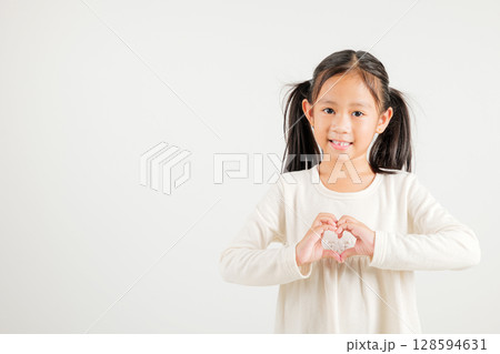 A smiling Asian young kid girl shows confidence, creating a heart shape with fingers and hands on white background. Portrait of kindergarten child sending love for Valentine's Day A smiling Asian young kid girl shows confidence, creating a heart shape with fingers and hands on white background. Portrait of kindergarten child sending love for Valentine's Day 128594631