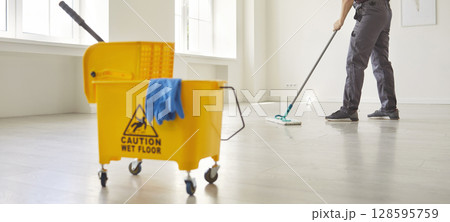 Janitor with mop cleaning empty office floor, worker mopping by cart with supplies and warning sign 128595759
