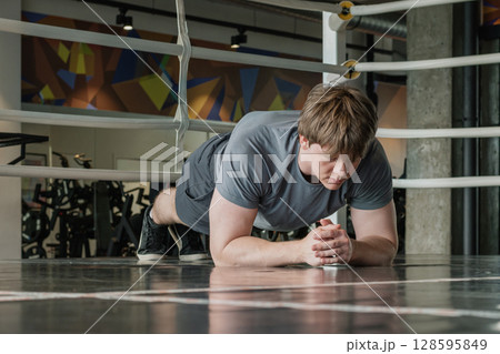 Young caucasian male person in gray sportswear does plank position at gym. Fitness center. Health care and workout. Young caucasian male person in gray sportswear does plank position at gym. Fitness center. Health care and workout. 128595849
