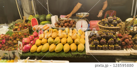 Colorful display of fresh tropical fruits at a night market in Thailand. Mangos, mangosteen, rambutan, dragon fruit and more arranged beautifully on a vibrant street stall. 128595880