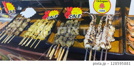 Grilled street food skewers in Thailand, including chicken satay, beef, octopus, and seafood, displayed on a market counter with price signs and vibrant labels. Grilled street food skewers in Thailand, including chicken satay, beef, octopus, and seafood, displayed on a market counter with price signs and vibrant labels. 128595881