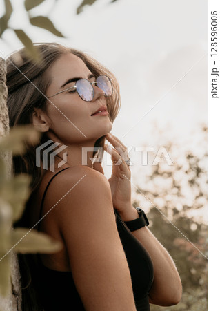 Woman Sunglasses Nature - Portrait of a young woman wearing sunglasses and looking away while standing next to a tree outdoors. Woman Sunglasses Nature - Portrait of a young woman wearing sunglasses and looking away while standing next to a tree outdoors. 128596006