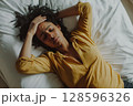 Woman With Curly Hair Lying on a Bed Looking Distressed and Holding Her Forehead in a Serene Room During Daytime 128596326