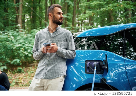 Trees behind. Handsome man is standing outdoors near electric automobile and waiting for charging 128596723