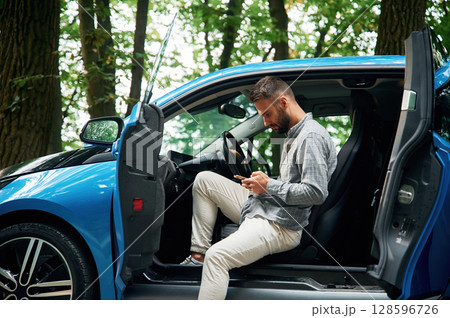 With smartphone in hands. Beautiful young man is sitting in the blue car outdoors With smartphone in hands. Beautiful young man is sitting in the blue car outdoors 128596726