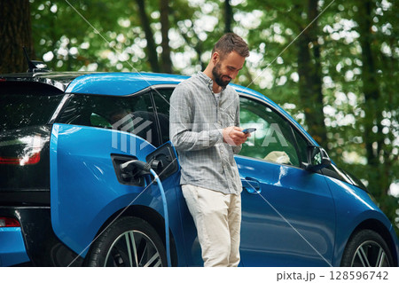 Wireless device, using smartphone. Handsome man is standing outdoors near electric automobile and waiting for charging Wireless device, using smartphone. Handsome man is standing outdoors near electric automobile and waiting for charging 128596742