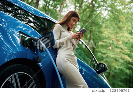 Young woman is standing outdoors near electric automobile that is charging 128596761