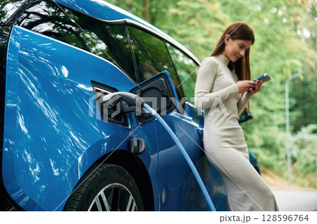 Leaning, using smartphone. Young woman is standing outdoors near electric automobile that is charging 128596764