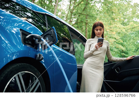 Leaning, using smartphone. Young woman is standing outdoors near electric automobile that is charging Leaning, using smartphone. Young woman is standing outdoors near electric automobile that is charging 128596766