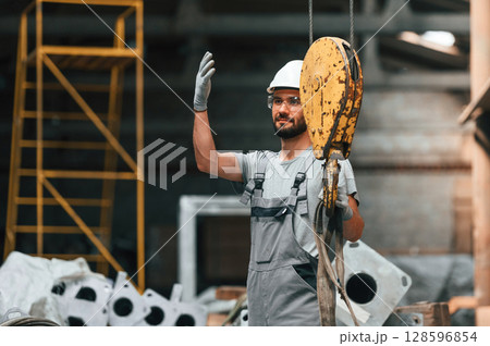 Using crane hook. Young factory worker in grey uniform Using crane hook. Young factory worker in grey uniform 128596854