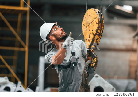 Yellow colored crane hook. Young factory worker in grey uniform Yellow colored crane hook. Young factory worker in grey uniform 128596861